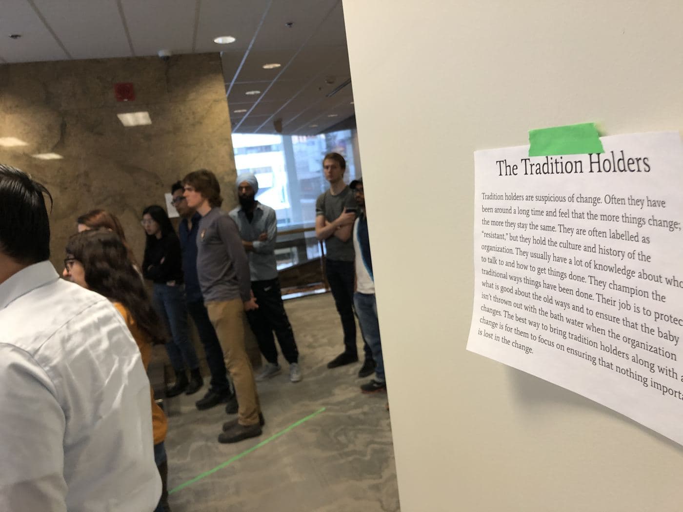 A printed description of the Tradition Holders taped to a wall while a group gathers beside it